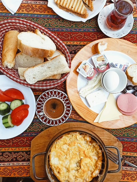 Kilim örtülü masada kahvaltı: ekmek sepeti, domates ve salatalık, peynir tabağı, çay ve sahanda menemen